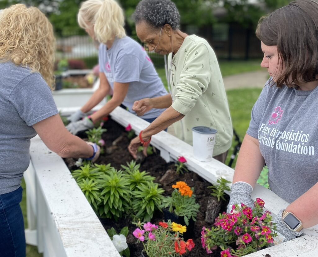 Four women are gardening together at Wesley Manor Retirement Community, planting colorful flowers in a raised white garden bed outdoors; they are wearing gloves and casual clothes, and trees are visible in the background.