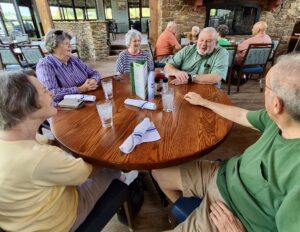 Five older adults sit around a wooden table at a restaurant, smiling and laughing together. Enjoying a warm, casual atmosphere, they celebrate the maintenance-free retirement lifestyle offered by Wesley Manor Independent Living.