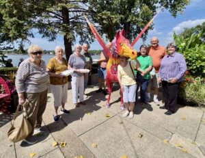A group of older adults stands outdoors on a sunny day, smiling around a colorful dragon sculpture at Wesley Manor Independent Living, enjoying maintenance-free retirement amid trees, plants, and a sparkling body of water in the background.