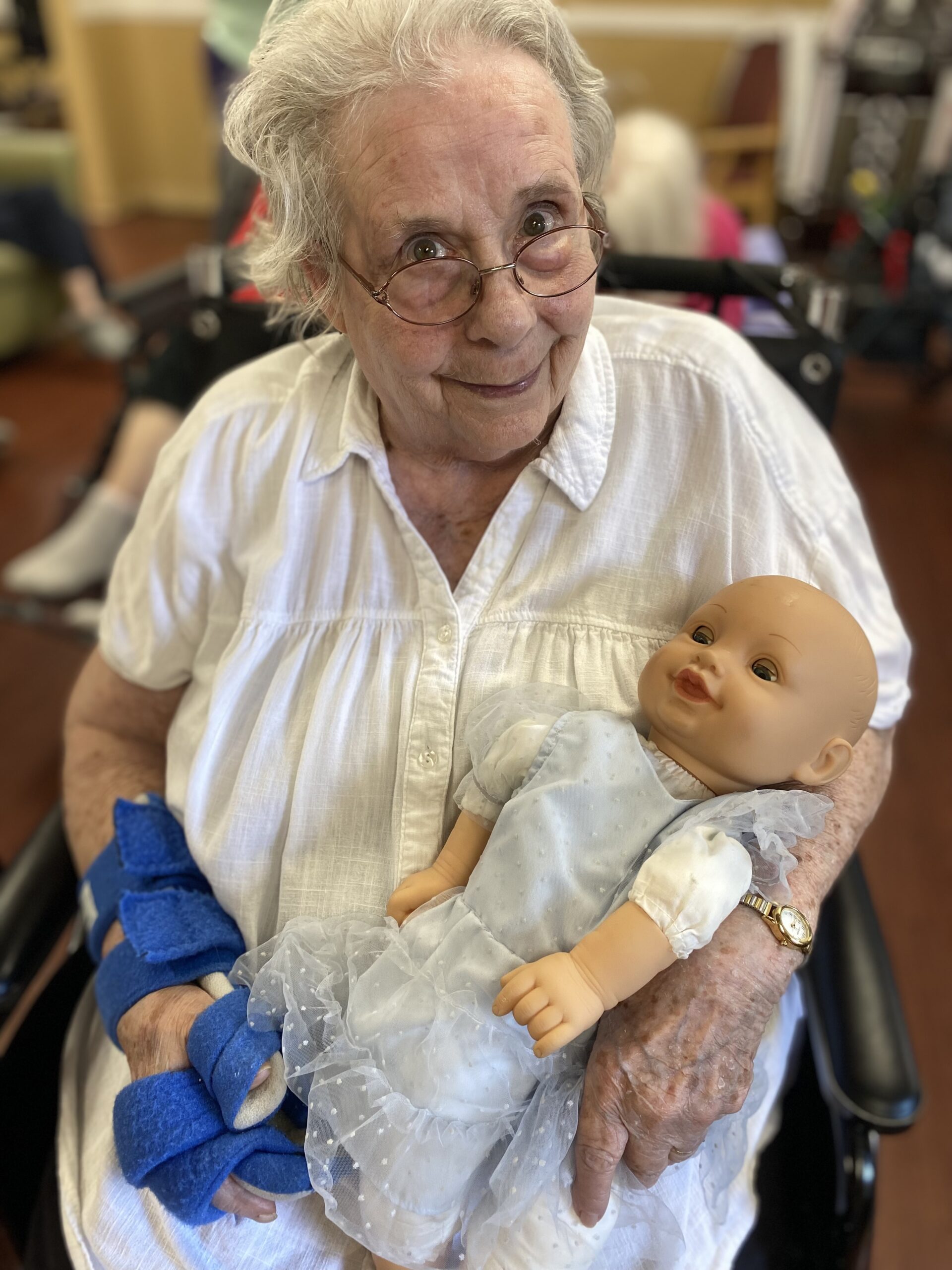 An elderly woman with glasses and a white blouse smiles while sitting in a wheelchair, holding a baby doll dressed in a light blue dress. She wears a blue brace on her left hand. The background shows blurred figures and furniture.