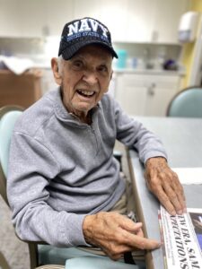 An elderly man in a “Navy” cap and gray sweater sits at a table, smiling and pointing at a newspaper. He is in a bright, clean room with light-colored cabinets and chairs.
