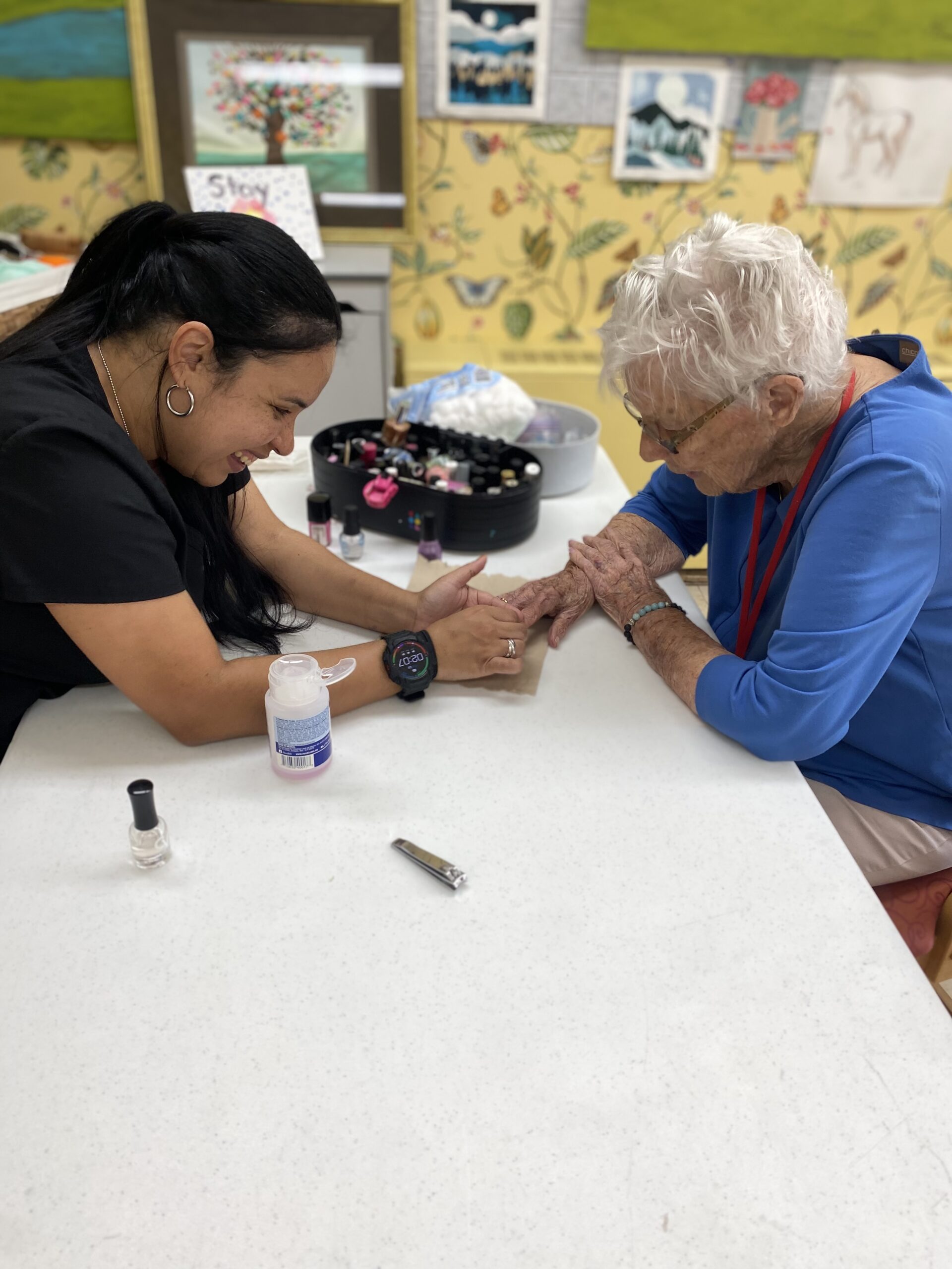 A smiling woman paints the nails of an woman sitting across from her at a table covered with nail polish bottles and manicure tools. Colorful art decorates the wall in the background.