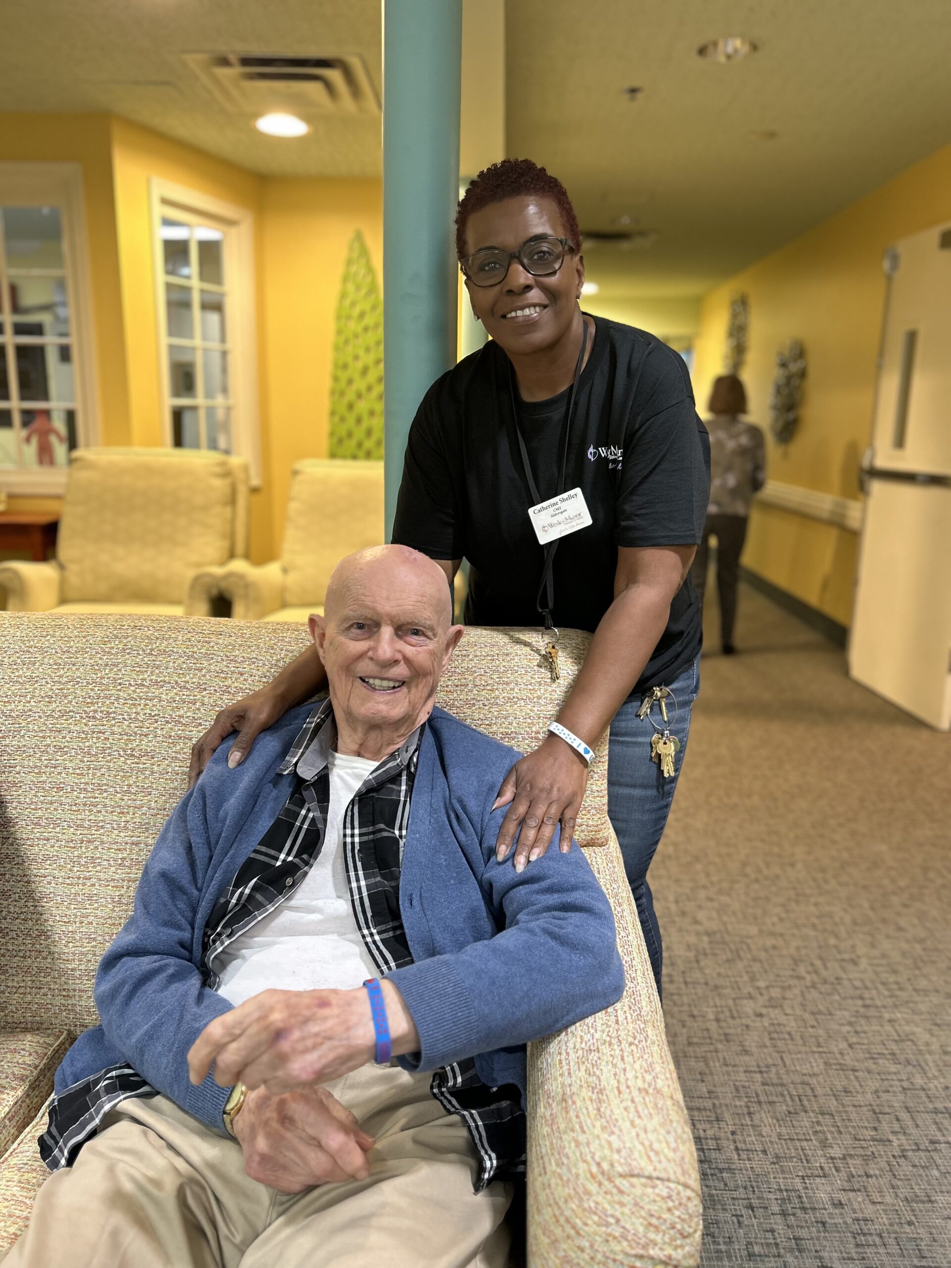 An elderly man sits smiling on a beige couch while a woman, standing behind him with her hands on his shoulders, smiles at the camera in a warmly lit room.