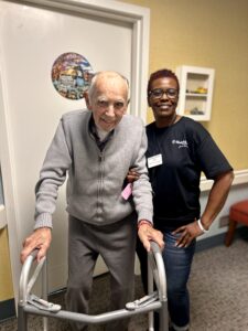 An elderly man using a walker stands smiling next to a woman in a black shirt and name tag who has her arm around his shoulder. They are indoors, in a hallway with a decorated door.