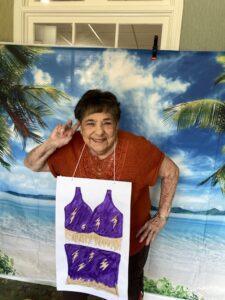 A woman smiles and flashes a peace sign, wearing an orange shirt and a paper cutout of a purple bikini over her clothes. She stands in front of a tropical beach backdrop with palm trees and blue sky.