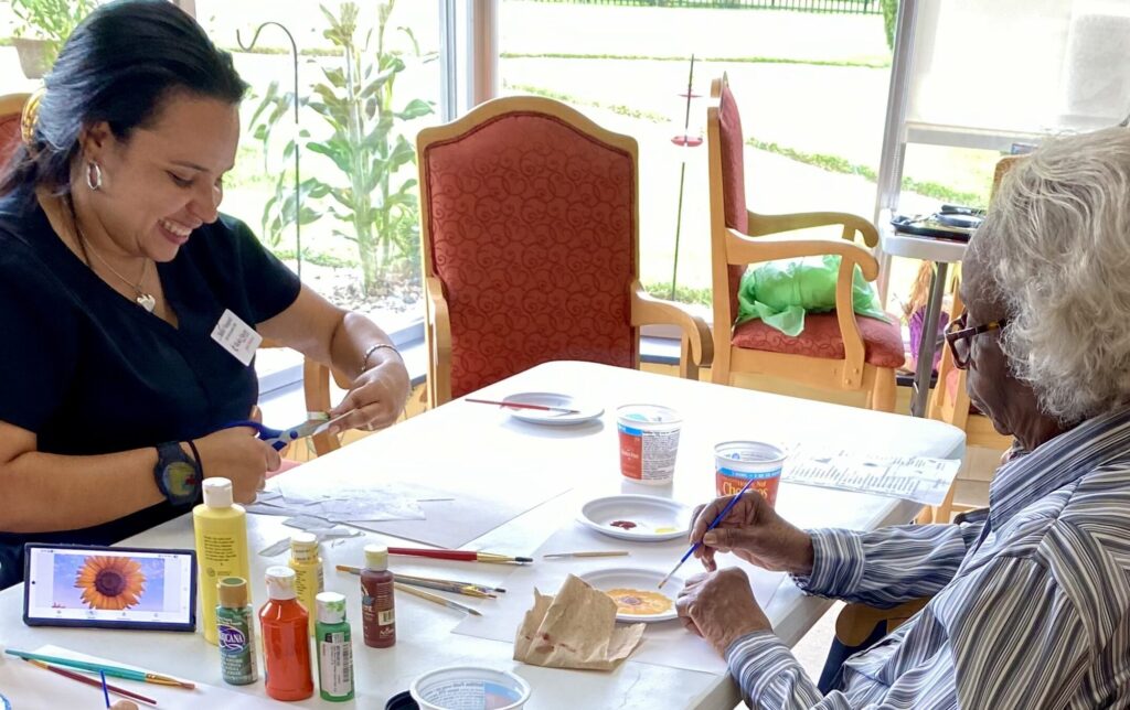 Two women enjoy art activities at a table filled with supplies, smiling and creating together. Bright natural light streams in, reflecting the vibrant community at Wesley Manor’s Personalized Senior Care & Assisted Living.