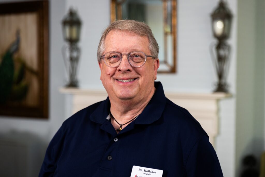A smiling older man with glasses and a navy blue polo shirt stands indoors. He wears a name tag and is posed in front of a white fireplace with decorative lanterns and a framed mirror.