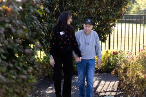 A memory care health professional and an elderly man hold hands and walk together outdoors along a garden path, surrounded by greenery and sunlight.