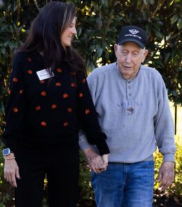 A memory care health professional and an elderly man hold hands and walk together outdoors along a garden path, surrounded by greenery and sunlight.