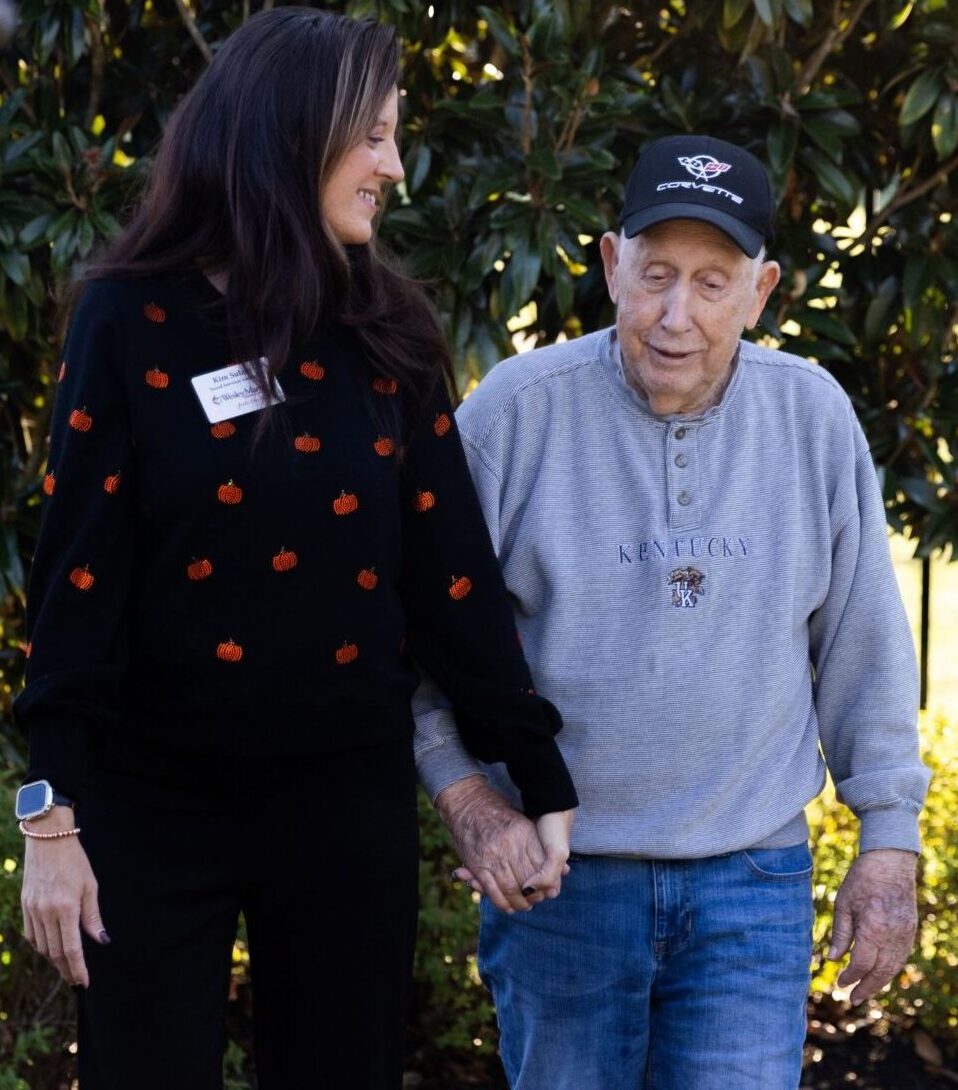 A memory care health professional and an elderly man hold hands and walk together outdoors along a garden path, surrounded by greenery and sunlight.