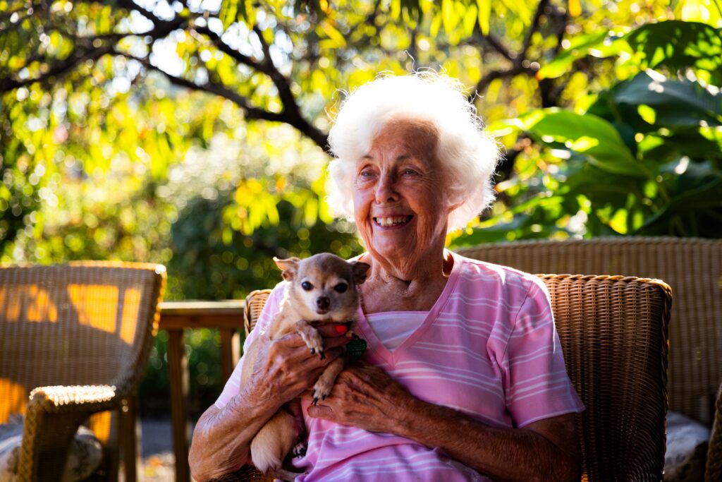 An elderly woman with white hair, wearing a pink striped shirt, sits on a wicker chair outdoors at a senior care facility specializing in memory care, smiling and holding a small chihuahua dog as sunlight filters through green foliage in the background.