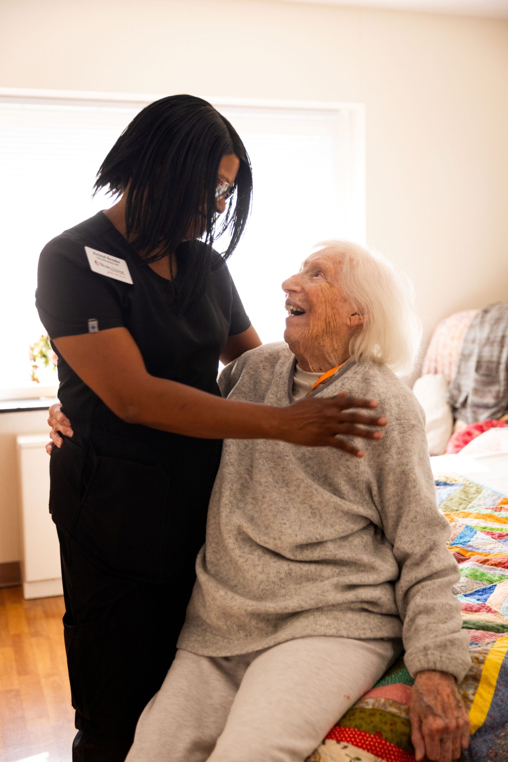 A memory care health professional in black scrubs smiles and embraces an elderly woman sitting on a bed. The elderly woman, in a gray sweater, looks up joyfully at the caregiver in a bright, cozy senior care facility.