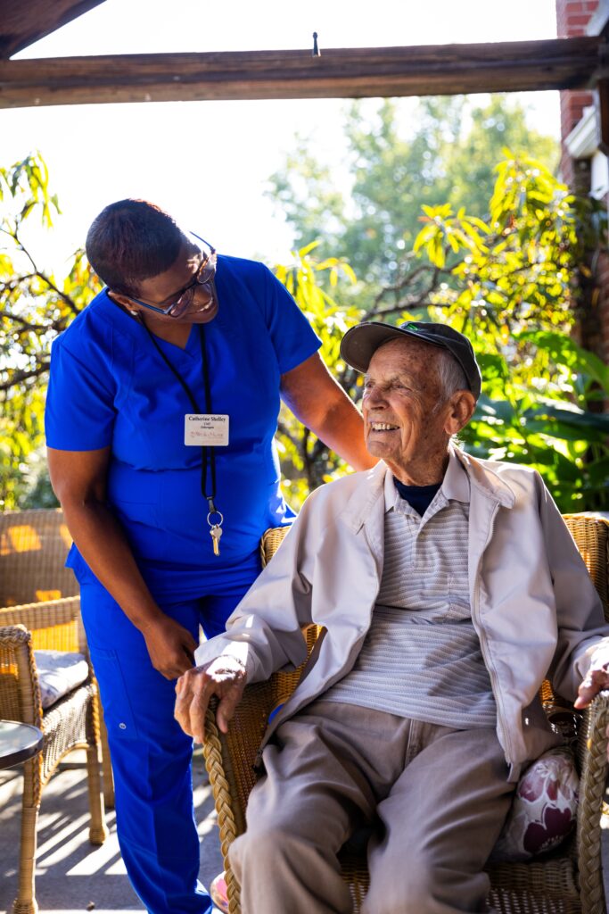 465A8174 A smiling elderly man sits outdoors in a wicker chair while a memory care health professional in blue scrubs stands beside him, kindly holding his hand. They appear to be enjoying a pleasant conversation in a sunlit garden at the senior care facility.