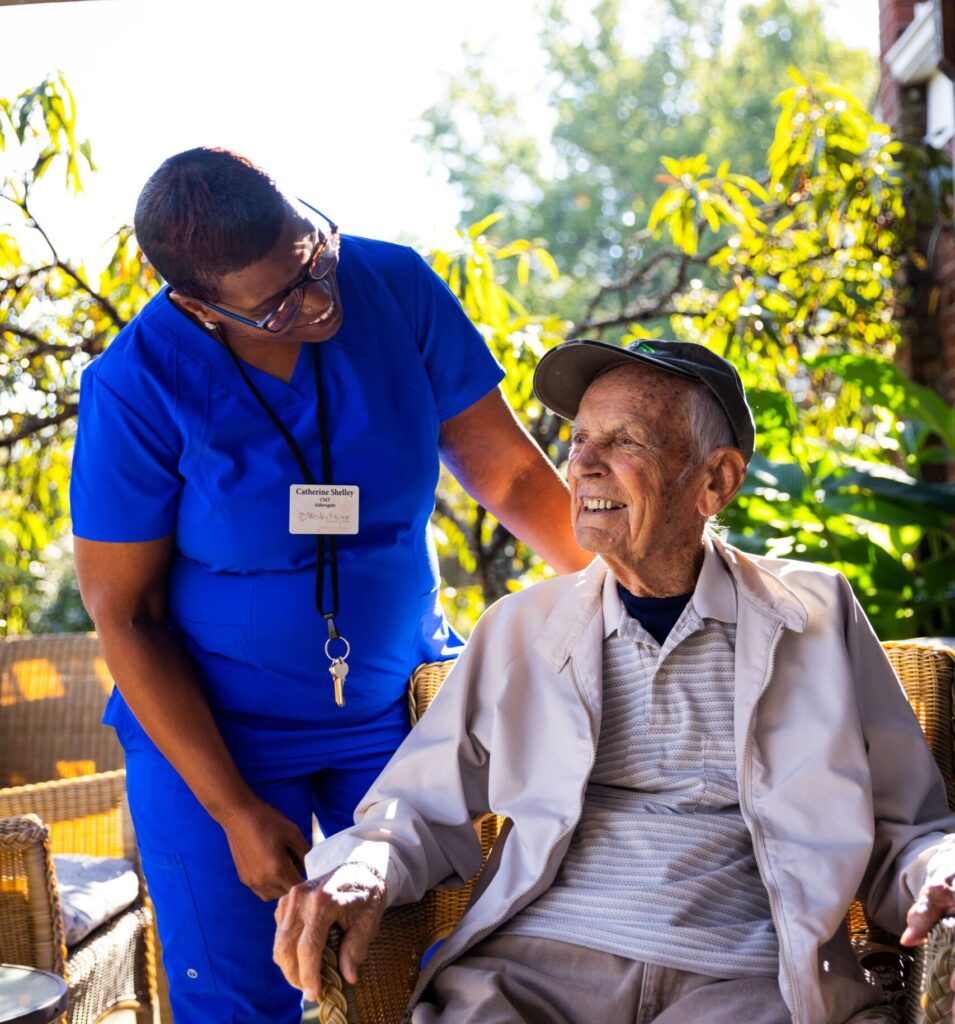 A smiling elderly man sits outdoors in a wicker chair while a memory care health professional in blue scrubs stands beside him, kindly holding his hand. They appear to be enjoying a pleasant conversation in a sunlit garden at the senior care facility.