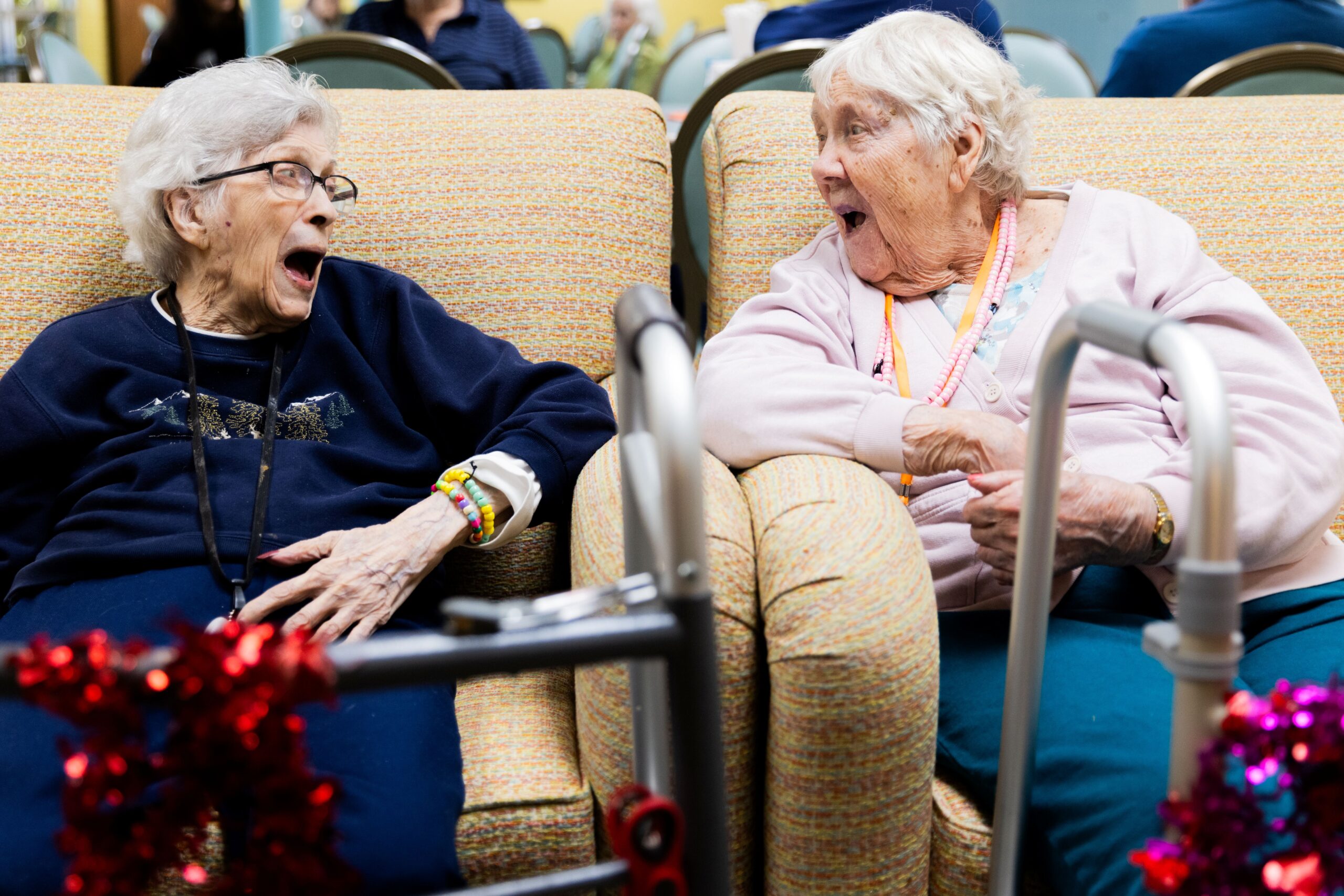 Two elderly women sit on sofas facing each other, laughing and smiling with mouths open wide at a senior care facility specializing in memory care. A walker and festive red garland are in the foreground, with others blurred in the background.