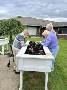 Three older adults tend to flowers in a raised white garden bed outside a senior care facility specializing in elderly care. Two women and one man work together, planting and caring for colorful flowers on a cloudy day.