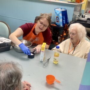 A memory care health professional in an orange apron measures ingredients at a table with two elderly women. Surrounded by cooking supplies, they appear to be baking together in a senior care facility specializing in elderly wellness.