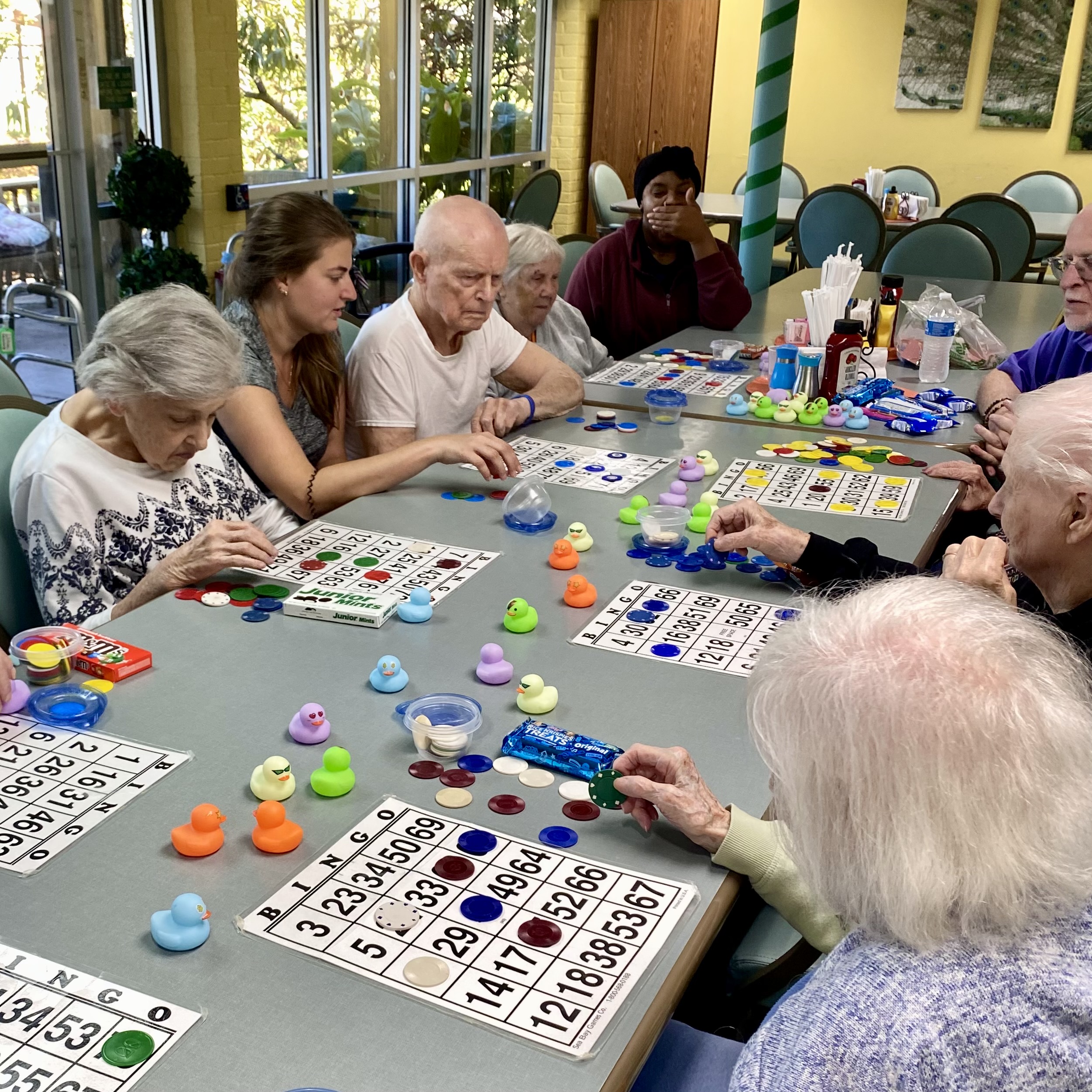 A group of elderly people and a memory care health professional sit around a table at a senior care facility, playing bingo with colorful chips and rubber ducks as markers. They appear focused and engaged, with bingo cards spread out before them.