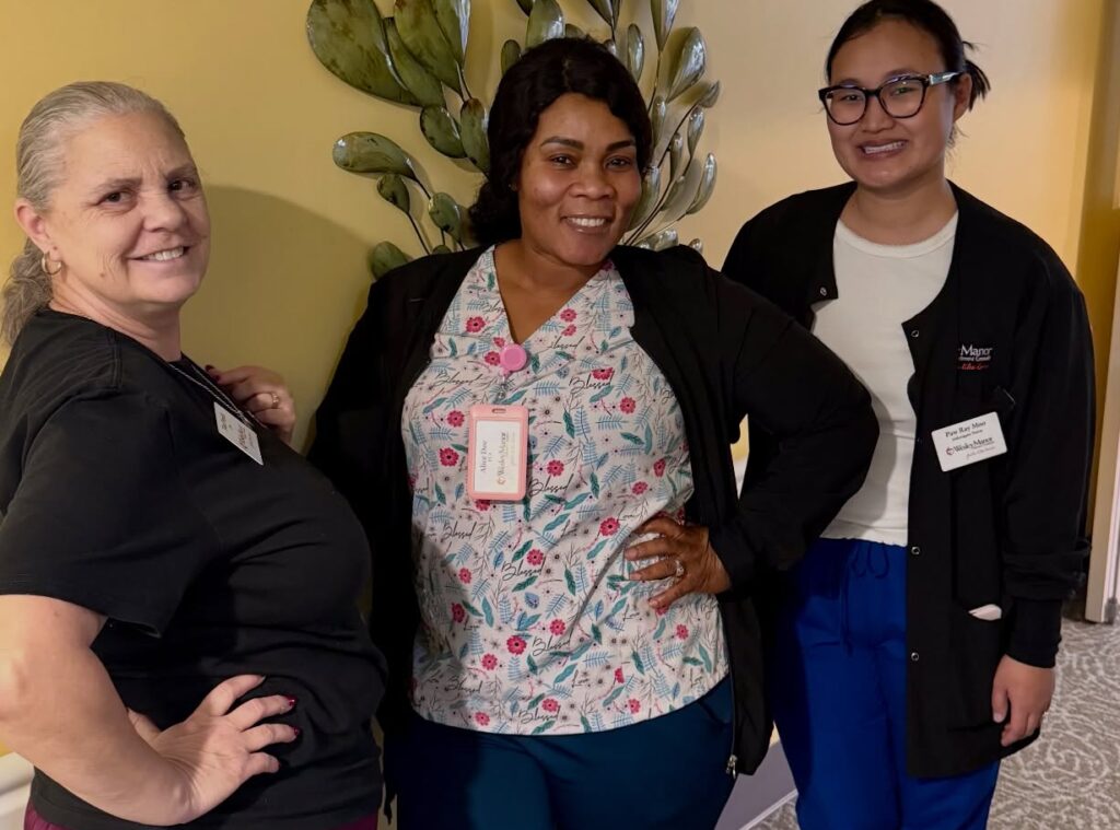 Three women in medical scrubs and name badges stand smiling together in a hallway with a yellow wall and leafy wall decor behind them.