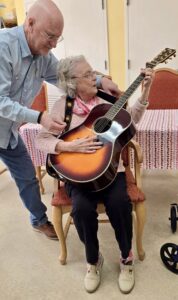 An elderly woman sits in a chair, holding an acoustic guitar, while an older man beside her helps position her hands—just one of the engaging moments at Assisted Living at Wesley Manor. A table with a striped cloth sits behind them indoors.