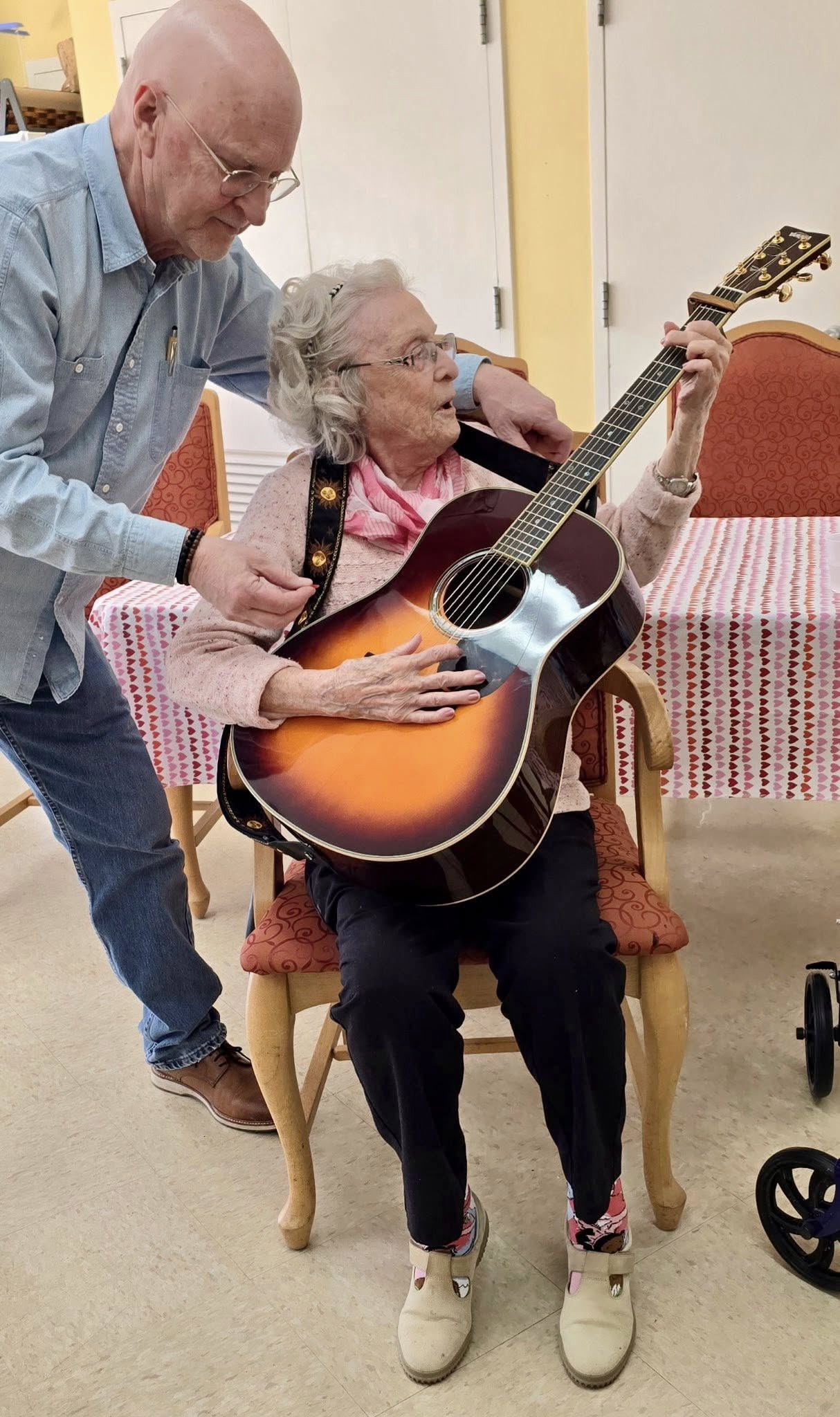 An elderly woman sits in a chair, holding an acoustic guitar, while an older man beside her helps position her hands—just one of the engaging moments at Assisted Living at Wesley Manor. A table with a striped cloth sits behind them indoors.