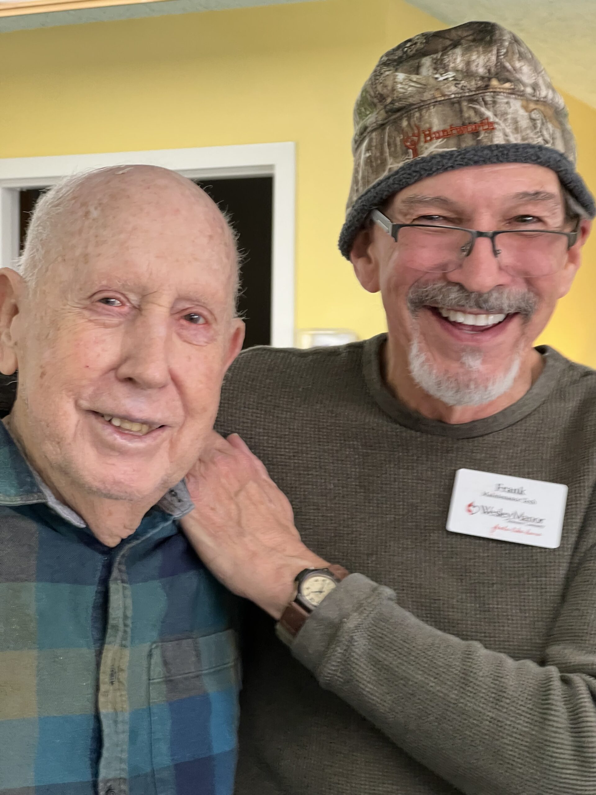 Two smiling older men pose together indoors. One wears a plaid shirt, and the other, with glasses and a beanie, has his arm around his friend’s shoulder by a yellow wall. Show your support for moments like these—donate to Wesley Manor.