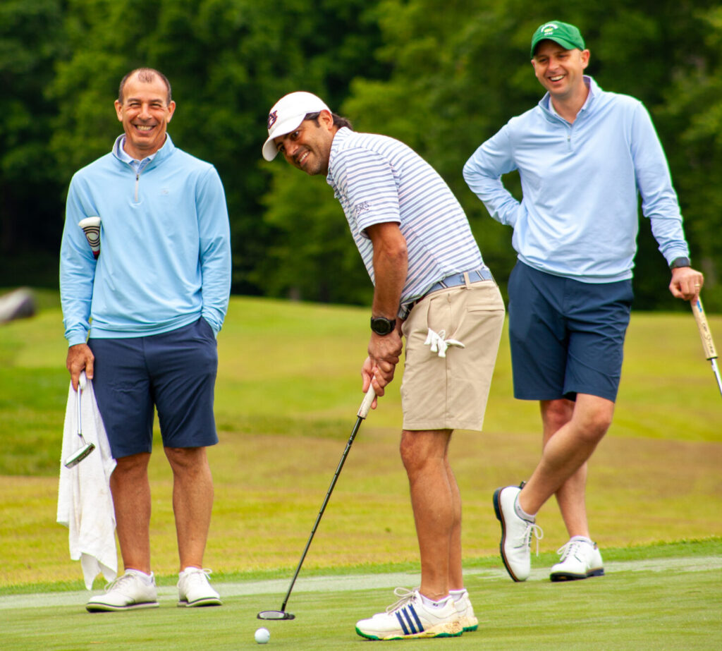 Three men stand on a golf course green during the Wesley Manor Swing for Seniors; one is putting while the other two, dressed in casual golf attire, watch and smile. Trees and lush greenery fill the background.