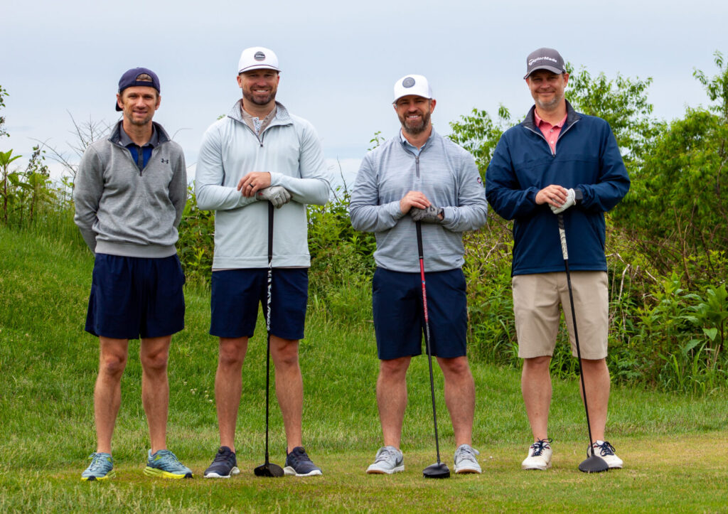 Four men stand side by side on a grassy golf course at the Wesley Manor Swing for Seniors, each holding a golf club. They are dressed in casual athletic clothing and hats, with greenery and a cloudy sky in the background.