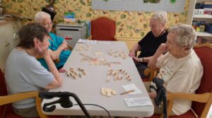 Four elderly women sit around a rectangular table in a rehab and skilled nursing facility, playing a tile game—possibly dominoes—in a cozy room with patterned wallpaper, bookshelves, and a walker nearby.