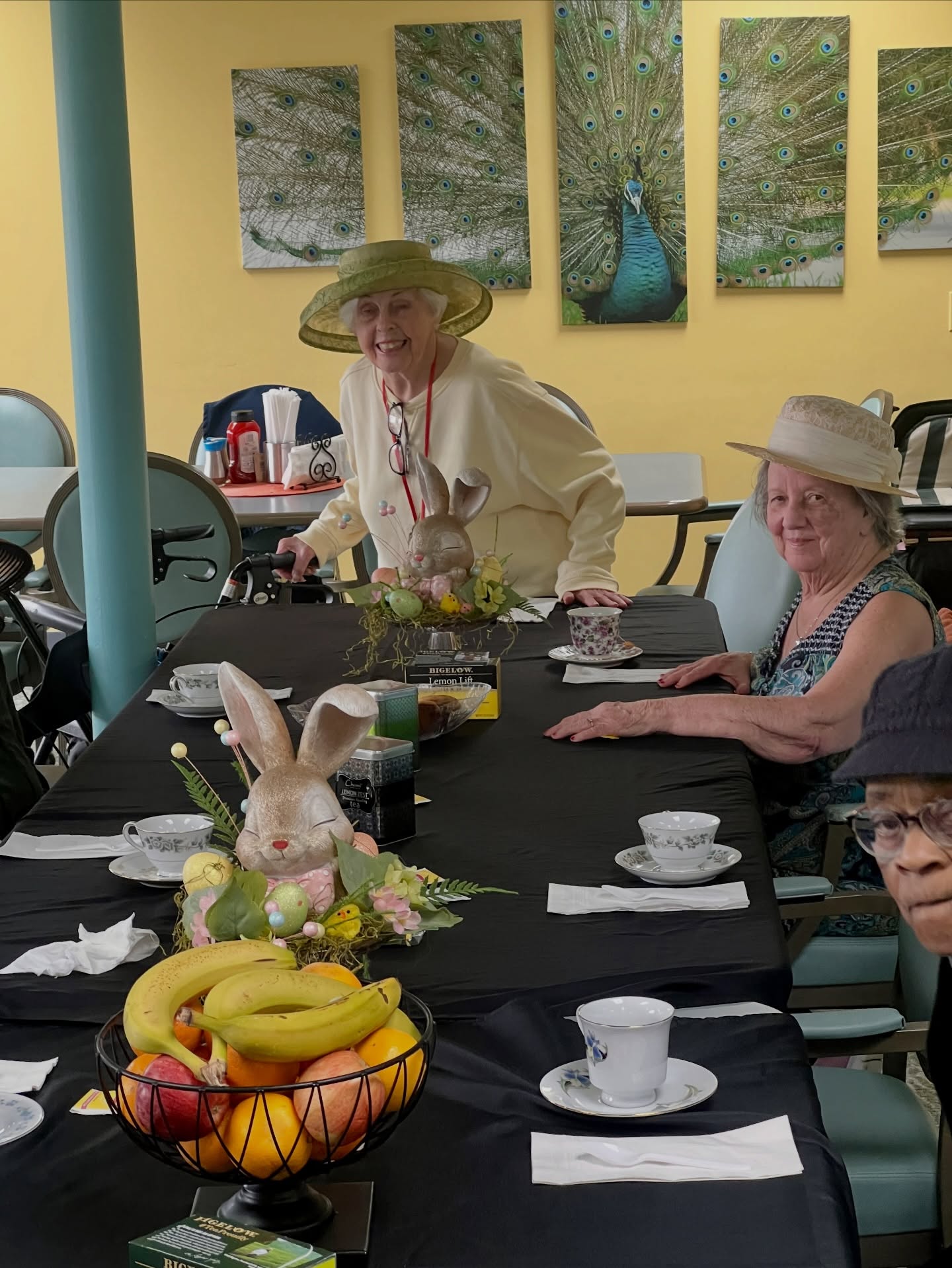 Two elderly women in hats sit at a long table with a black tablecloth, decorated with fruit, rabbit figurines, and teacups. Peacock artwork hangs on a yellow wall behind them in the cozy Wesley Manor memory care setting.