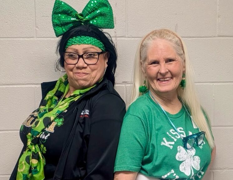 Two women dressed in green outfits stand back-to-back and smile. One wears a large green bow, scarf, and headband, while the other wears shamrock earrings and a shirt that says “Kiss Me.” Both pose against a light brick wall.