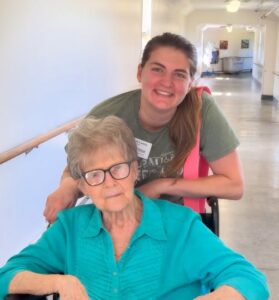 A smiling woman stands behind an older woman seated in a wheelchair in a brightly lit rehab and skilled nursing hallway. The older woman wears glasses and a teal blouse; the younger woman wears a green shirt and a name tag.