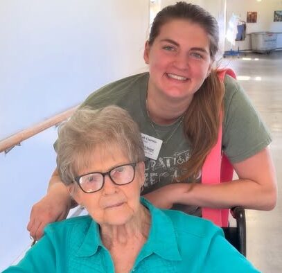 A smiling woman stands behind an older woman seated in a wheelchair in a brightly lit rehab and skilled nursing hallway. The older woman wears glasses and a teal blouse; the younger woman wears a green shirt and a name tag.