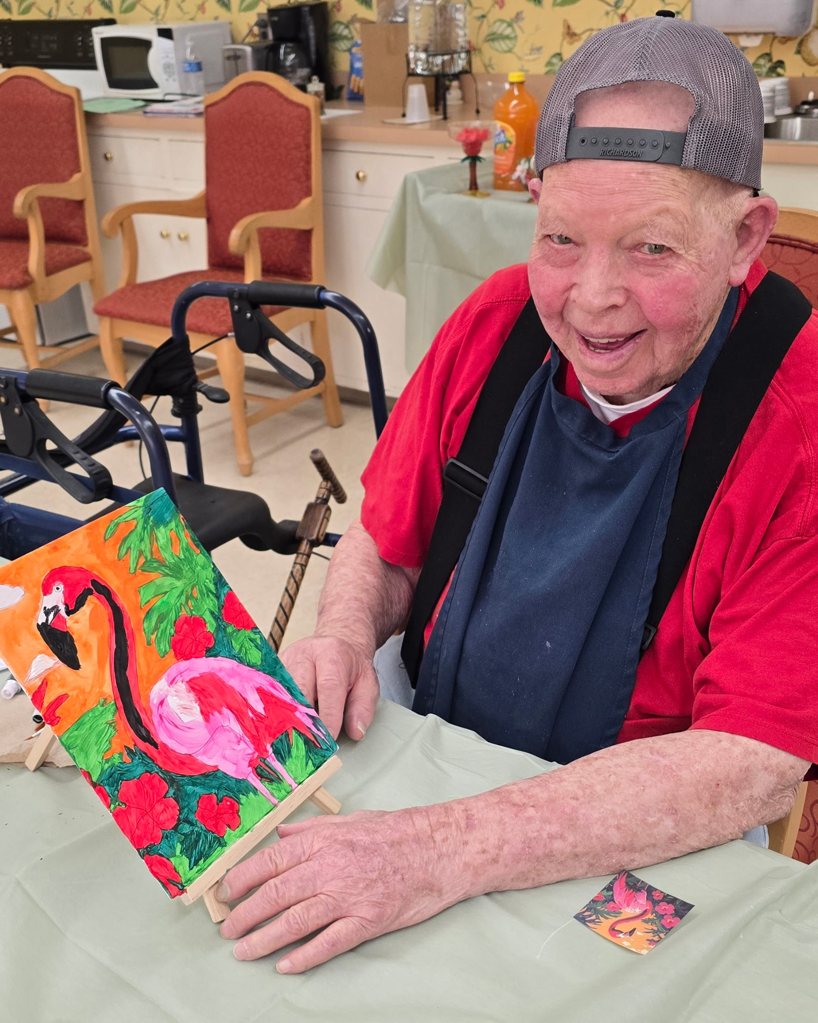 An elderly man at Assisted Living at Wesley Manor, in a red shirt and backwards cap, smiles while holding a colorful flamingo painting at a table in a bright room with art supplies and chairs in the background.