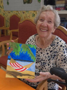 An elderly woman with short gray hair, wearing a polka dot shirt, smiles while holding up a colorful painting of a hammock on a beach—reflecting the creative and vibrant Wesley Manor lifestyle.