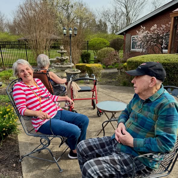 Two older adults sit and chat in a garden patio at Wesley Manor memory care, with one woman smiling in a striped shirt and a man in a cap and plaid shirt. Nearby, another person sits with a red walker amid trees and shrubs.