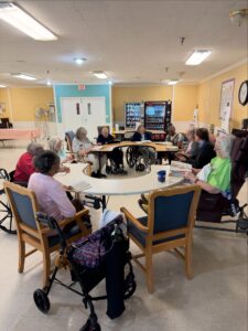 A group of elderly adults, enjoying the Wesley Manor lifestyle, sit in a circle at a care facility—some in wheelchairs, others in chairs—engaged in a group activity or discussion. Tables and a vending machine can be seen in the background.