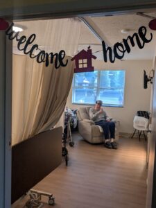 A welcome home sign hangs above a doorway to a room where an elderly person sits in a recliner near a window, with a hospital bed and medical equipment visible nearby.