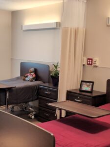 A hospital or care facility room with two beds, one with a red cover and the other with a stuffed animal. There is a nightstand with a plant and a framed photo, and a privacy curtain partially drawn between the beds.