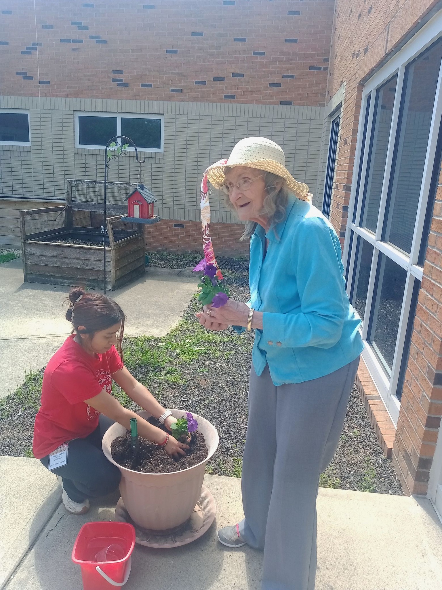 An elderly woman in a sunhat holds a flower while a young woman kneels beside a large pot, planting flowers together in the sunlit courtyard garden at Assisted Living at Wesley Manor.