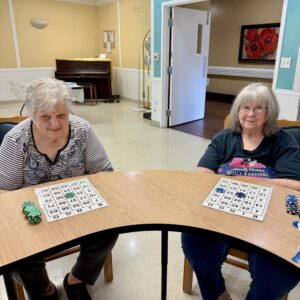 Two older women sit at a table in a rehab and skilled nursing facility, playing bingo with cards and poker chips. They are indoors in a brightly lit room with a piano and art on the walls in the background.