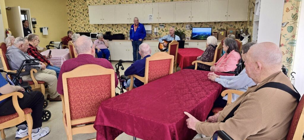 A group of elderly people sit around tables in a room, listening to a man standing and another playing guitar. The room has floral wallpaper, a TV, and cabinets in the background. Some people use wheelchairs or walkers.