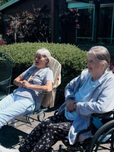 Two older women sit outside on a sunny day at Assisted Living at Wesley Manor, one in a wheelchair and the other in a patio chair. They appear relaxed and happy, enjoying the fresh air amid green bushes and a building in the background.