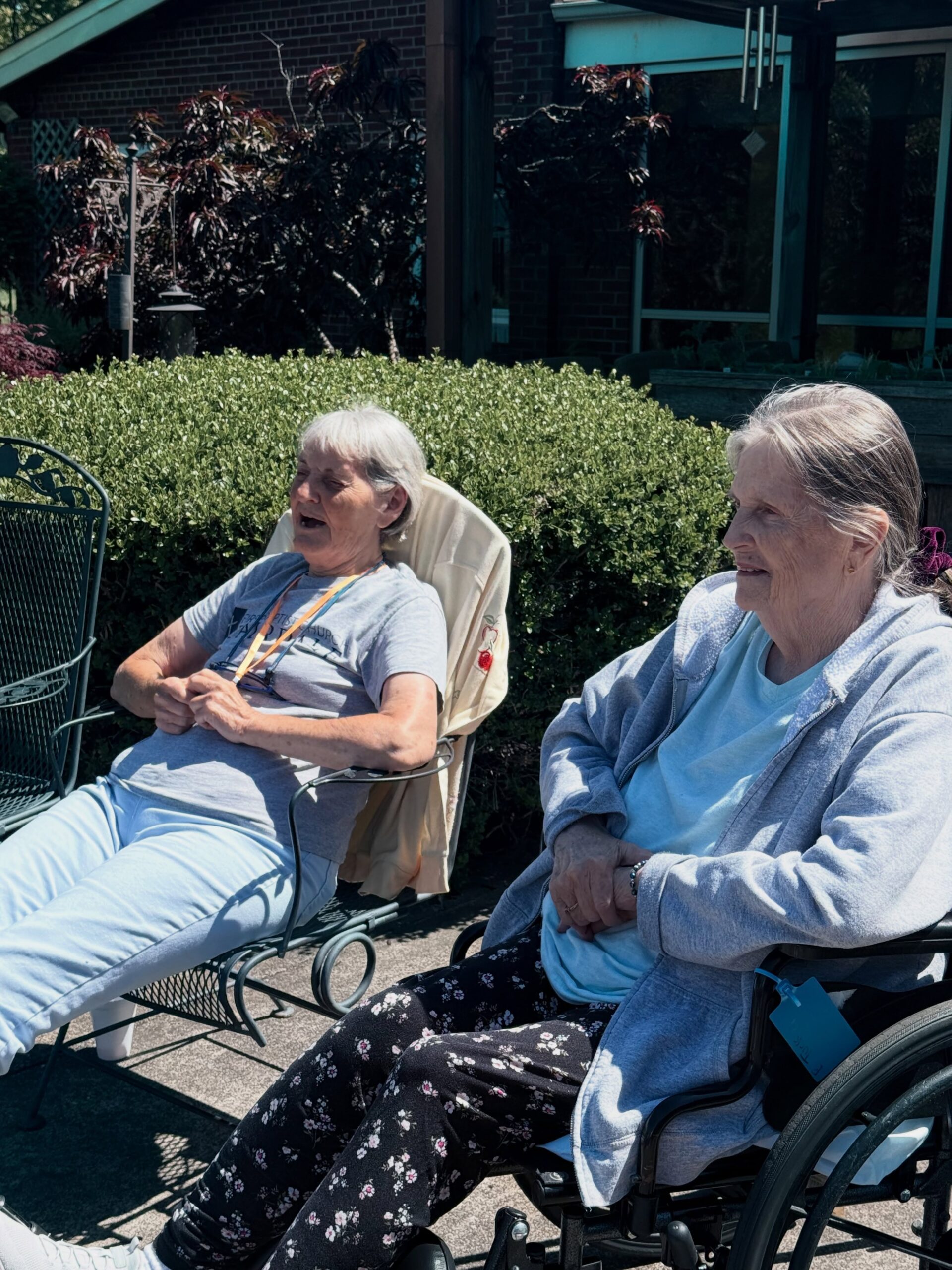 Two older women sit outside on a sunny day at Assisted Living at Wesley Manor, one in a wheelchair and the other in a patio chair. They appear relaxed and happy, enjoying the fresh air amid green bushes and a building in the background.
