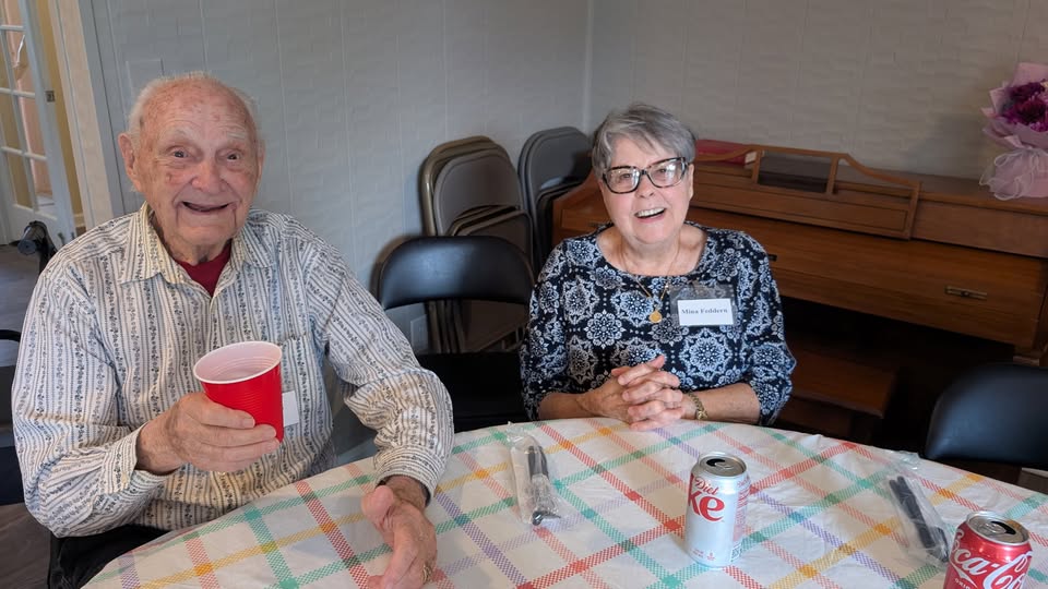An elderly man holding a red cup and a woman with glasses and a name tag sit at a table with a checkered tablecloth, Diet Coke cans, and folded utensils, smiling at the camera.