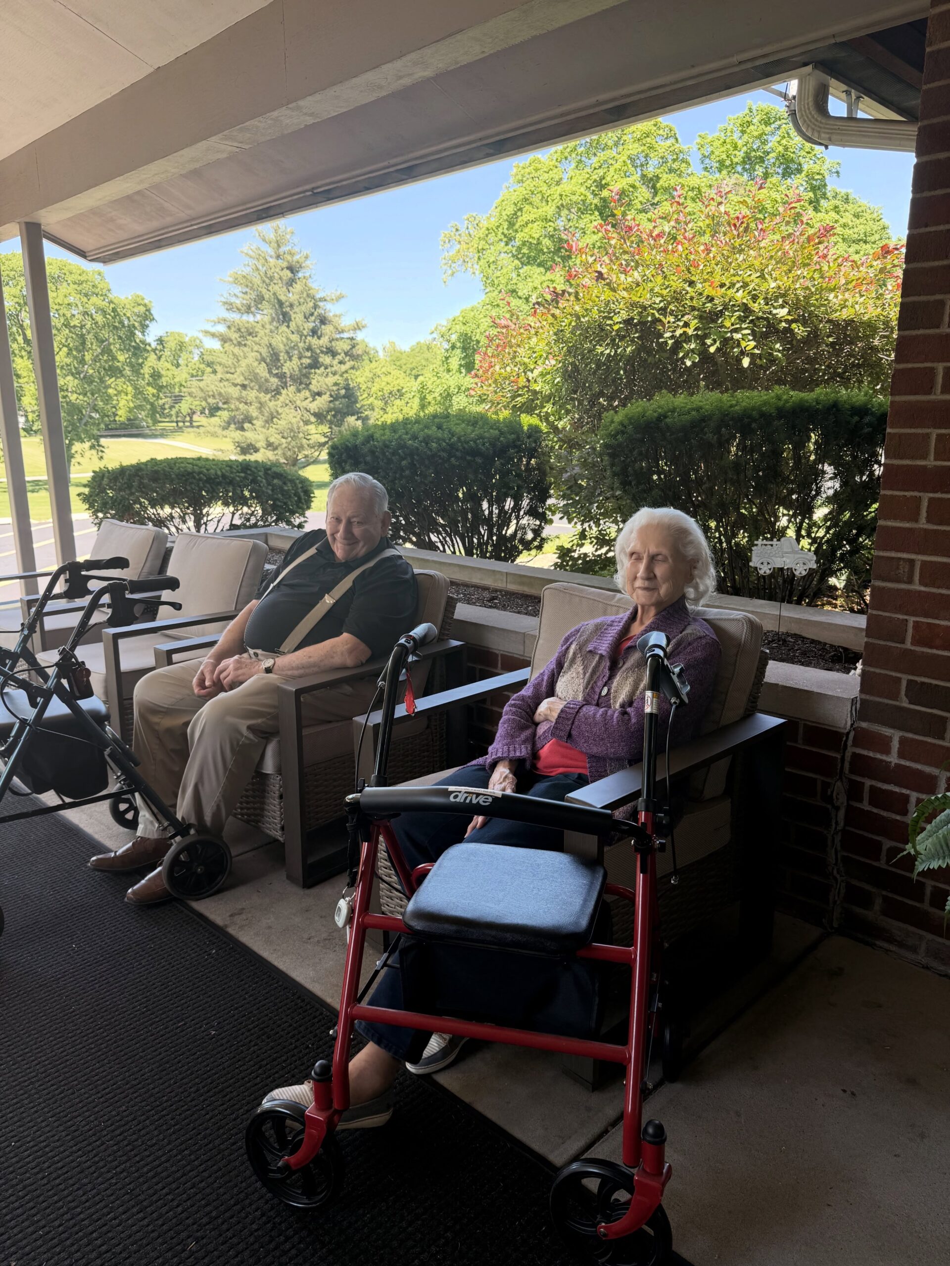 An elderly man and woman sit outside a brick building at Assisted Living at Wesley Manor, each with a walker nearby. They relax under a covered patio, surrounded by greenery on a bright, sunny day.