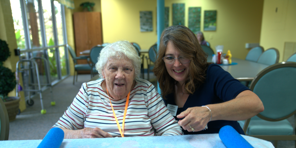 An elderly woman and a younger woman smile together at a table in a brightly lit room, capturing the cheerful spirit often found at Wesley Manor. Discover more moments like this in our Wesley Manor senior living FAQs.