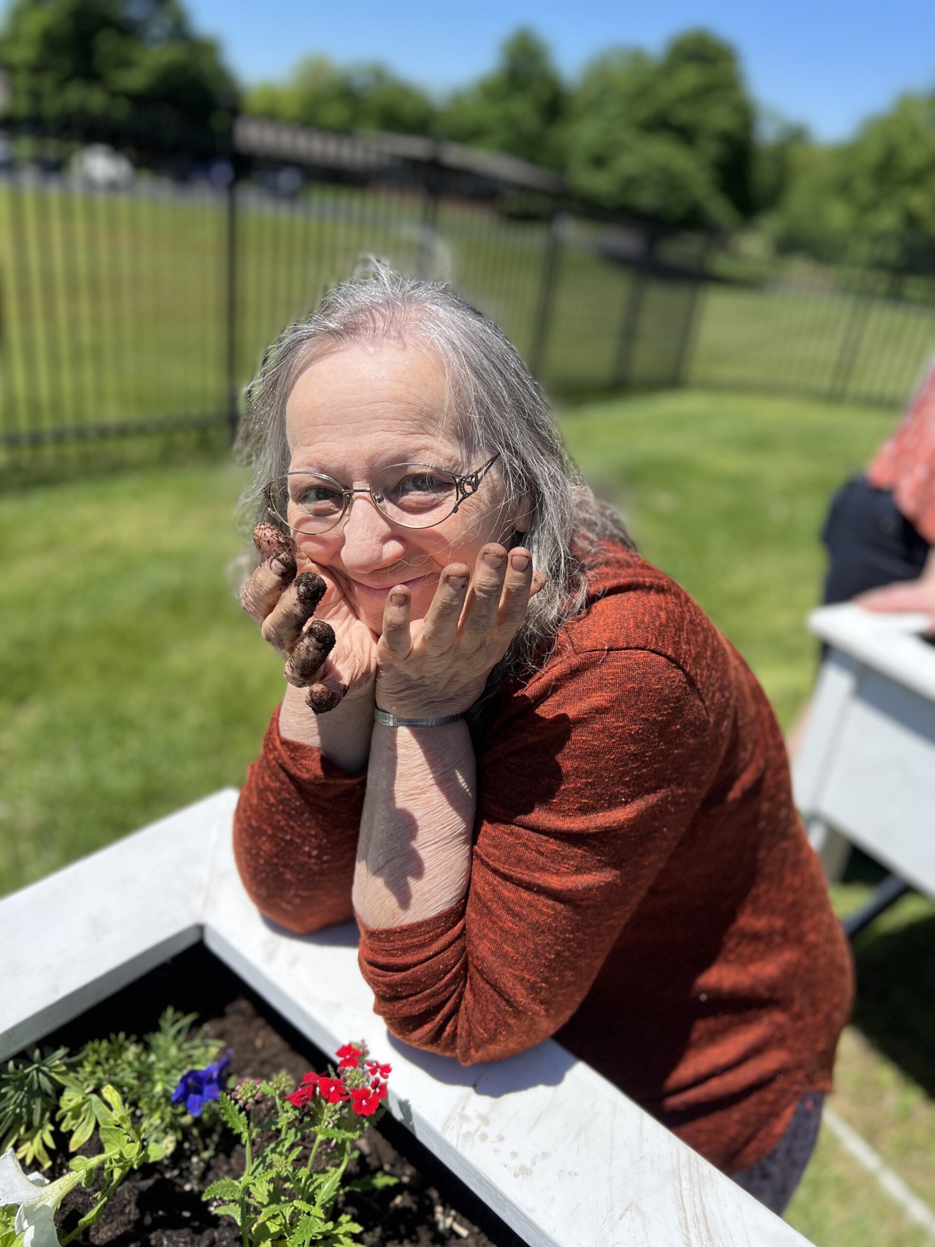 An older woman with gray hair and glasses, wearing an orange shirt, smiles playfully with her chin resting on her muddy hands beside a raised garden bed at a rehab and skilled nursing facility on a sunny day.