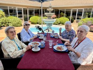 Four elderly women sit around a table with meals and drinks, smiling outdoors by a fountain under a blue parasol, enjoying a sunny day at Assisted Living at Wesley Manor.