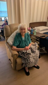 An elderly woman with white hair, wearing a green blouse and black shoes, sits smiling in a recliner with a patterned blanket on her lap. A bed and medical supplies are nearby, and another person is in the background.
